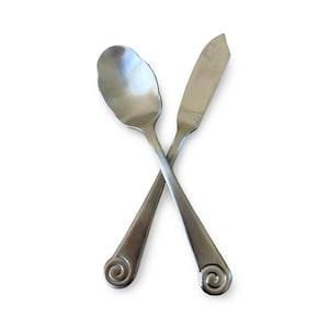 May include: A stainless steel spoon and butter knife are crossed against a white background. The spoon has a scalloped bowl, and both utensils have a decorative swirl at the end of their handles. The image is well-lit and shows the details of the silverware.