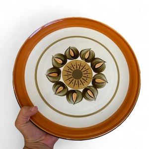 Puede incluir: Un plato redondo de cerámica con borde marrón y centro blanco. El plato presenta un diseño floral en tonos marrón, verde y beige. El plato tiene aproximadamente 25 cm de diámetro.