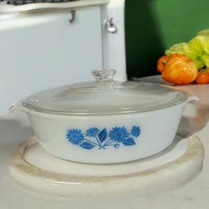 Puede incluir: Una fuente blanca para horno con tapa de cristal transparente. El plato presenta un estampado floral azul en los laterales. El plato está sobre una superficie redonda, blanquecina, similar al mármol. La tapa tiene un pomo de cristal transparente.