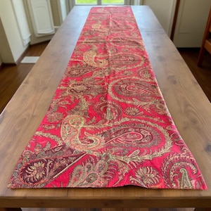 May include: A red paisley table runner with a repeating pattern of large, ornate paisley designs in shades of gold, green, and burgundy. The runner is rectangular and lies on a wooden table.
