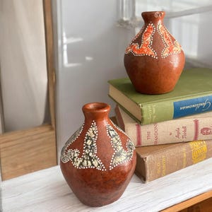 May include: Two brown ceramic vases with decorative patterns in white, black, and orange. The vases have a bulbous shape and a narrow neck. They are displayed on a white surface with books in the background.