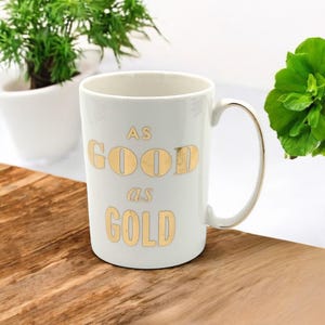 以下が含まれることがあります： 「AS GOOD AS GOLD」と金色の文字が入った白いセラミックマグ。 マグには金色のハンドルとトリムが付いています。 マグは、白い背景と緑のある木製の表面にあります。