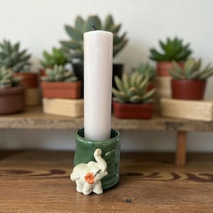 Może przedstawiać: Biała świeca kolumnowa w zielonym ceramicznym świeczniku z białą figurką słonia. Uchwyt ma teksturowany wzór. Sukulenty w terakotowych donicach znajdują się w tle na drewnianej półce.