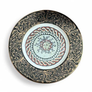 Puede incluir: Plato decorativo con borde negro con filigrana dorada y centro de mosaico. El mosaico representa una cara similar al sol con detalles en rojo, blanco y negro. El plato tiene un borde dorado.