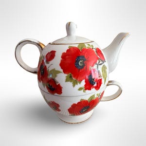 May include: White ceramic teapot and teacup set decorated with red poppy flowers and green leaves. The teapot has a lid and a gold-trimmed handle and spout. The teacup is stacked beneath the teapot.