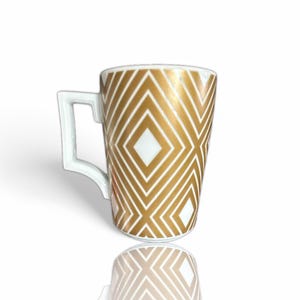 Può includere: Una tazza in ceramica bianca con un motivo geometrico a rombi dorati. La tazza ha un manico bianco e una finitura lucida. Il design è simmetrico e moderno, per caffè o tè.