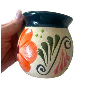May include: A hand-painted ceramic mug with a cream-colored base and a dark teal rim. The mug is decorated with a large orange flower, green leaves, pink accents, and black swirls. The mug is held by a hand.