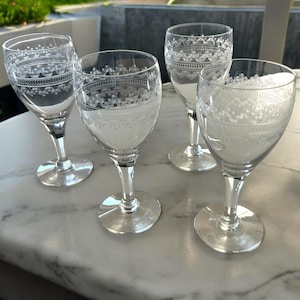 May include: Four clear wine glasses with etched floral patterns. The glasses have a delicate design around the rim and a textured pattern on the bowl. The stems are slender, and the bases are round. The glasses are arranged on a marble surface.