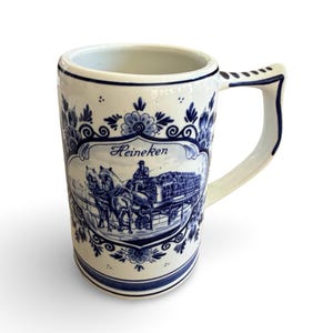 May include: A white ceramic beer stein with blue floral and scenic designs. The stein features the word "Heineken" and an illustration of a horse-drawn carriage. The handle is curved and the rim is trimmed with blue.