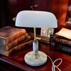 May include: A white banker's lamp with a curved shade, brass accents, and a marble base. The lamp is on a wooden desk next to a stack of antique books. The lamp's design is classic and elegant.