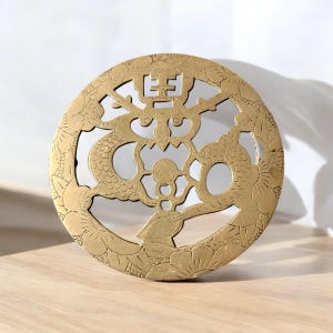 May include: A round, gold-colored metal decorative piece with a dragon and floral design. The intricate cutout pattern features a central Chinese character. The piece is likely used for decorative purposes.