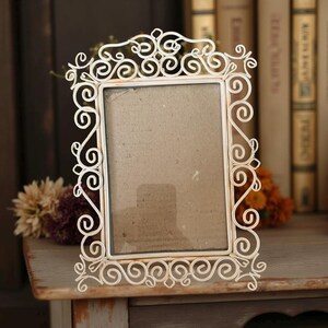 May include: A cream-colored, ornate metal picture frame with a rectangular opening. The frame features intricate scrollwork and stands on a wooden surface. The background includes books and flowers, suggesting a vintage aesthetic.