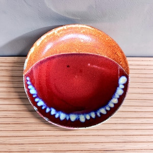 May include: A round ceramic plate featuring a two-tone glaze. The upper section is a textured, speckled orange, while the lower portion is a deep, glossy red with a decorative border of blue and cream-colored drips along the bottom edge.