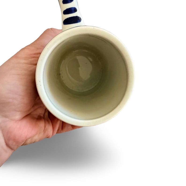 May include: A white ceramic mug with a blue handle. The handle features three blue oval shapes. The inside of the mug is a light green color. The mug is held in a person's hand.