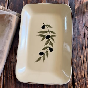 May include: A rectangular cream-colored ceramic serving tray featuring a painted olive branch motif in the center. The design includes dark black olives and muted green leaves. The edges of the tray have a distressed brown finish.