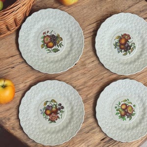 May include: Four white ceramic plates with a scalloped edge and a fruit design in the center. The plates are arranged on a wooden surface, with a basket of fruit and loose fruit nearby. The fruit design includes pears, grapes, and apples.