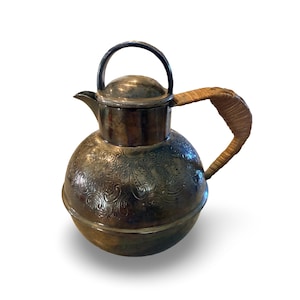 May include: A silver-colored, vintage-style tea or coffee pot with a round body and a lid. The pot features an etched floral design and a woven handle. The handle is made of a light brown material. The pot has a spout for pouring.