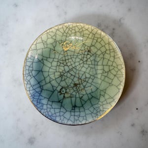 May include: A small, round ceramic dish with a crackle glaze pattern in shades of blue and green. The dish has a gold rim and delicate gold accents. The overall aesthetic is reminiscent of antique Asian pottery.