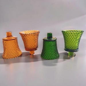 May include: Four glass votive candle holders featuring a hobnail texture. Two holders are amber-colored, one is a deep forest green, and one is a bright lime green. Each piece has a flared rim and a small, cylindrical base for stability.