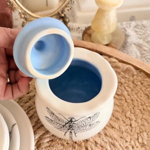 May include: A small, off-white ceramic jar with a blue interior and lid. The jar features a black dragonfly illustration on the front. The lid is light blue. The jar is open, revealing the blue interior. Decorative item.
