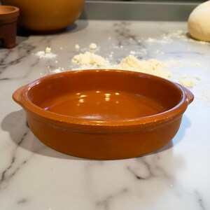 Könnte beinhalten: Eine runde, terrakottafarbene Keramik-Backform mit zwei kleinen Griffen. Die Form ist leer und steht auf einer Marmorarbeitsplatte. Weitere Keramikschalen und Mehl befinden sich im Hintergrund.