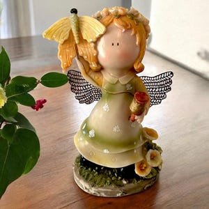 Puede incluir: Figura de un pequeño ángel con cabello naranja, vestido verde y alas negras. El ángel sostiene una mariposa amarilla y un carrete de hilo. La base está decorada con flores y vegetación.