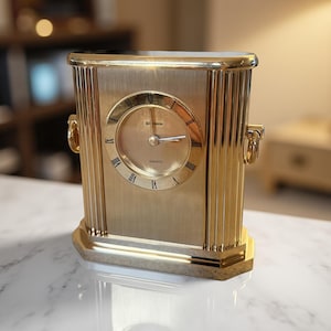 May include: A gold-toned quartz clock with a rectangular body and a round clock face. The clock features Roman numerals and a brushed metal finish. The clock has a decorative design with vertical lines and ring handles on the sides.