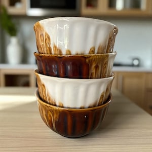 Puede incluir: Una pila de cuatro cuencos de cerámica con un diseño con volantes. Los cuencos alternan entre blanco y marrón, con un efecto de esmalte goteado. Los cuencos están apilados sobre una superficie de madera.