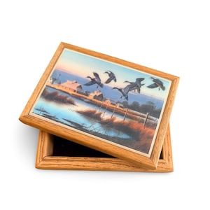 May include: Wooden box with a hinged lid. The lid features a painting of ducks in flight over a marshy landscape with buildings in the background. The box is made of light-colored wood and has a rectangular shape.