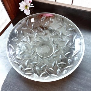 May include: A clear glass serving bowl with a leaf design. The bowl is round with a slightly raised rim and a central circular indentation. The leaf pattern radiates from the center. The bowl is likely used for serving salads or fruit.