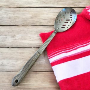 Puede incluir: Una espumadera de acero inoxidable con mango de madera descansa sobre un paño de cocina a rayas rojas y blancas. La cuchara tiene una cabeza ovalada con agujeros y un mango de madera remachado. El paño está doblado sobre una superficie de madera clara.