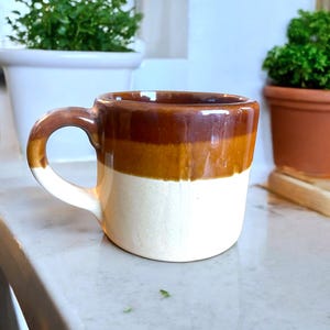 May include: A ceramic mug in brown, tan, and white. The mug has a rounded shape with a handle, suitable for drinks. It is placed on a white surface. The mug's design is simple and classic.