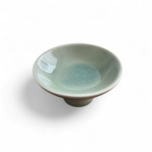 May include: A small, celadon-colored ceramic bowl with a pedestal base. The bowl has a crackle glaze and a slightly darker blue-green hue in the center. The bowl is round and has a shallow depth, suitable for serving small portions.