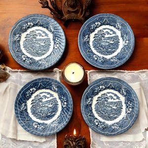 May include: Four blue and white decorative plates with a landscape design. Each plate features a central image of a building and trees, surrounded by a floral border. A lit candle sits in the center, with a white cloth underneath.