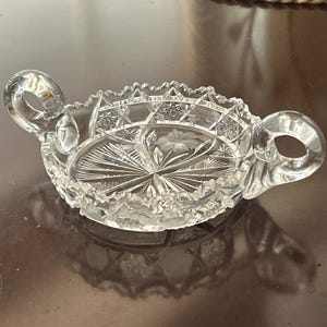 Puede incluir: Un plato de cristal tallado transparente con dos asas de bucle. El plato presenta intrincados patrones de estrellas y flores, con un borde festoneado. El diseño es simétrico, con una flor grabada en el centro. El plato es probablemente vintage.