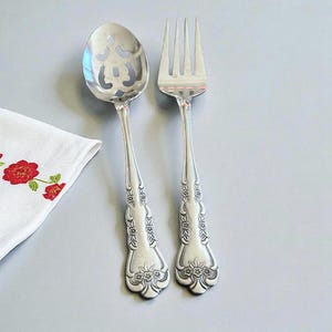 May include: Silverware set featuring a slotted spoon and a fork. Both pieces have ornate floral detailing on the handles. A white napkin with red floral embroidery is in the corner. The silverware is likely for serving.