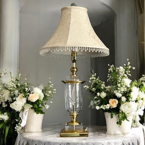 May include: A vintage table lamp with a beige patterned lampshade and a clear glass base with a wheat design. The lamp has a gold-colored metal base and accents. The lamp is surrounded by white flowers in white vases.