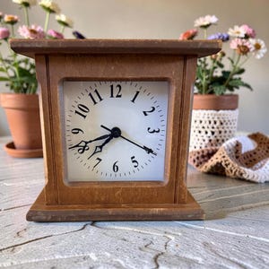 May include: A wooden square-faced desk clock with a white clock face and black hands and numbers. The clock is brown and sits on a flat surface. The clock is surrounded by flowers in pots.