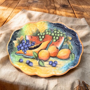May include: A hand-painted ceramic serving platter with a scalloped edge. The platter features a colorful still life of fruit, including watermelon, cantaloupe, grapes, blueberries, and oranges. The background is a mix of yellow, green, and blue.