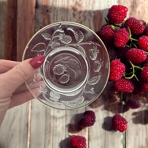 Puede incluir: Un platillo de vidrio transparente con borde dorado y diseño de frambuesas en relieve. Frambuesas rojas frescas están esparcidas alrededor del platillo sobre una superficie de madera. El platillo es sostenido por una persona.