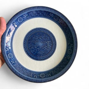 Puede incluir: Un plato redondo de cerámica blanca y azul con una detallada escena de paisaje en el centro. El plato presenta un borde azul con patrones florales y geométricos. El plato está sostenido por una mano.