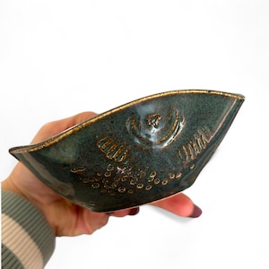 以下が含まれることがあります： 魚のデザインが施された、手作りのティール色のセラミックボウル。 ボウルはユニークな形状で、魚のエンボス加工が施され、質感のある表面と金茶色の縁があります。 ボウルは白い背景に対して手で持たれています。