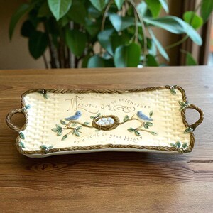 Puede incluir: Bandeja rectangular de cerámica con un patrón tejido y bordes en forma de cuerda. Presenta una rama con dos pájaros azules, un nido con huevos y el texto "May your day be brightened by a song in your heart."