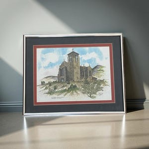 May include: Framed watercolor print of a stone church on a hill, under a blue sky with clouds. The artwork is framed in silver with a black and red mat. The church has a tall steeple and several windows.