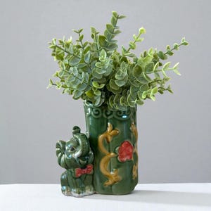 May include: A green ceramic vase with an elephant figurine attached. The vase is filled with green leafy stems. The vase has gold and red floral accents. The elephant has a red bow tie.