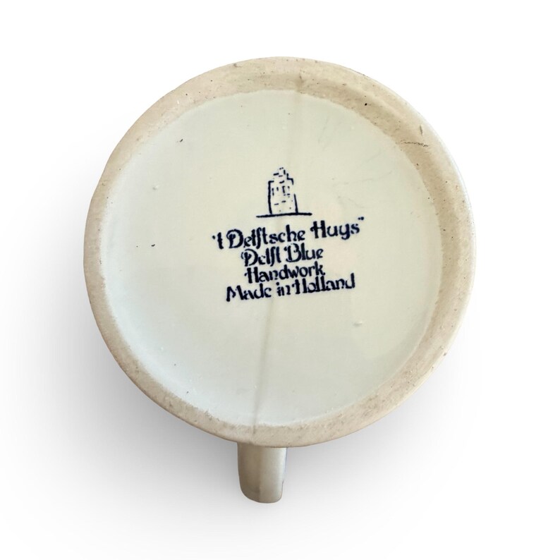 May include: A white ceramic mug with a light beige handle. The bottom of the mug features blue text that reads "'Delftsche Huys' Delft Blue Handwork Made in Holland" and a small illustration of a building.