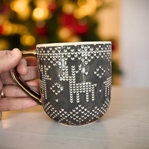 May include: A dark gray ceramic mug with a gold handle and rim. The mug features a white pattern of deer and other shapes. The mug is held by a hand, with a blurred background of Christmas lights.