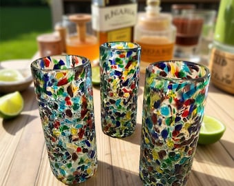 May include: Three colorful, speckled glass tumblers. The glasses are cylindrical with a textured surface, showcasing a mosaic of red, blue, green, and yellow glass pieces. Limes and bottles are in the background, suggesting a bar or drink setting.