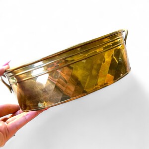 May include: A rectangular brass planter with a geometric design. It features two small handles at each end. The brass has a shiny, reflective surface. The planter is held by a person.