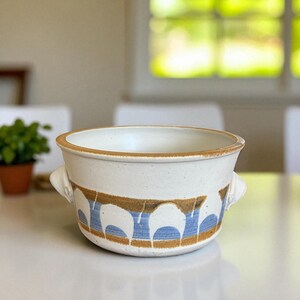 May include: A ceramic bowl with a cream-colored exterior and a brown rim. The bowl features a decorative band of blue and brown arches around the middle and has two small handles. This handcrafted bowl is suitable for serving.